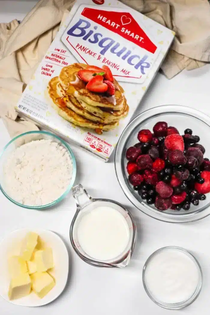 A box of Bisquick pancake mix is surrounded by bowls containing flour, mixed berries perfect for a blackberry cobbler, a cup of milk, a dish of butter, and a container of sugar. The ingredients are arranged on a white surface with a beige cloth in the background.