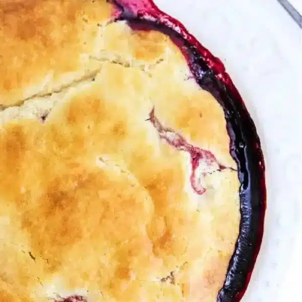 A close-up of a freshly baked bisquick pie with a golden-brown crust, slightly cracked, revealing a glimpse of dark red filling beneath, reminiscent of a blackberry cobbler. A silver spoon rests on a white plate beside the pie.