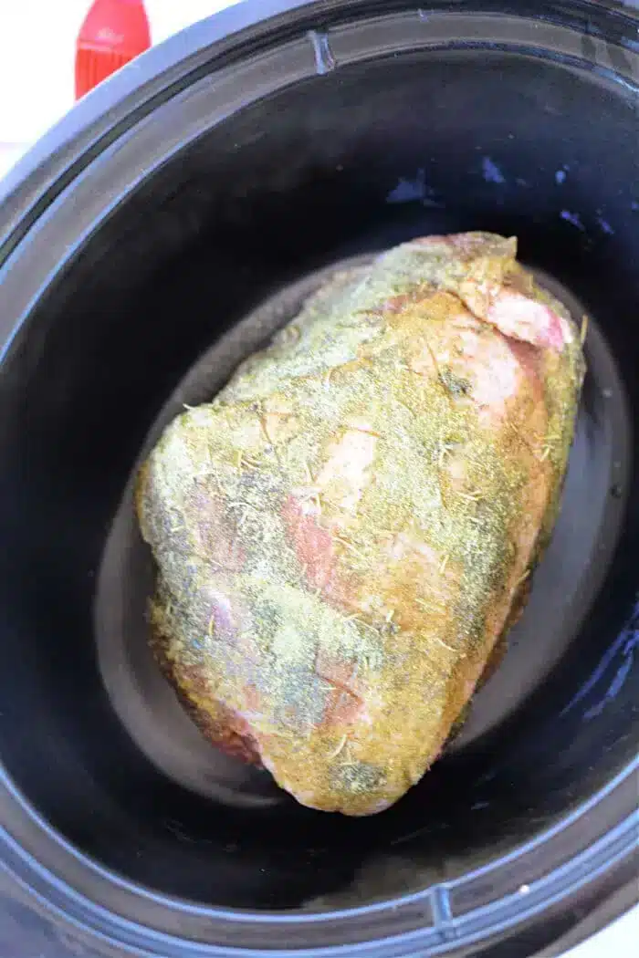 A seasoned piece of meat is placed inside a slow cooker. The 3 Ingredient Pork Roast is covered in herbs and spices, ready to be cooked.