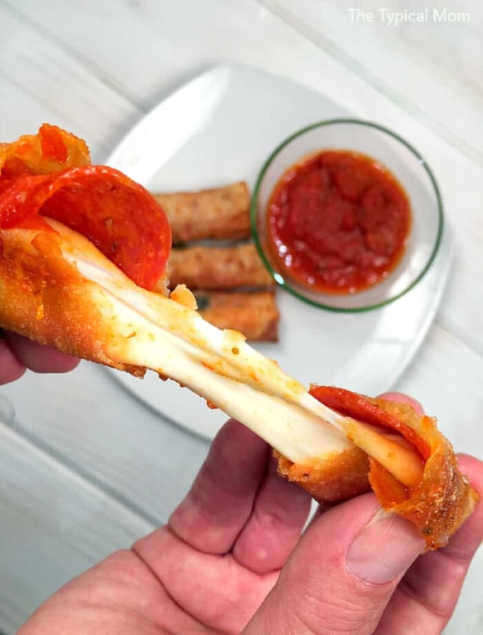 A hand holds a cheesy pepperoni pizza stick made with egg roll wrappers, with sauce and more sticks on a plate in the background—one of the tastiest recipes using egg roll wrappers.