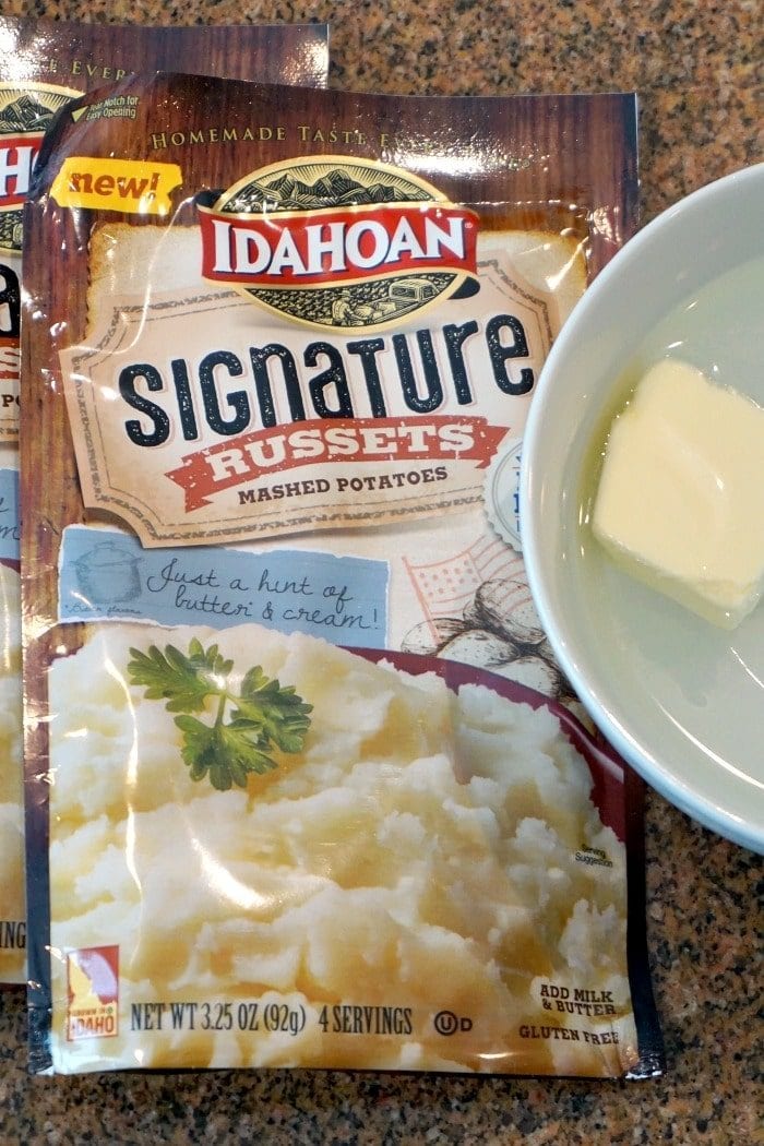 A package of Idahoan Signature Russets mashed potatoes sits invitingly next to a bowl with a pat of butter, perfect for crafting a delicious Shepherds Pie with instant potatoes.