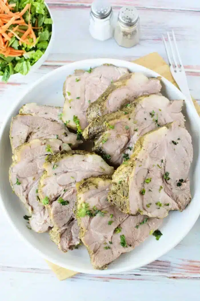 A white plate holds several slices of 3 Ingredient Pork Roast Slow Cooker-style cooked pork, garnished with chopped herbs.