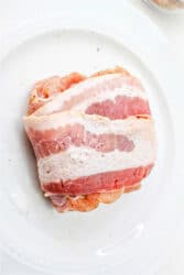 Raw bacon-wrapped chicken thighs rest on a white plate, ready for cooking.