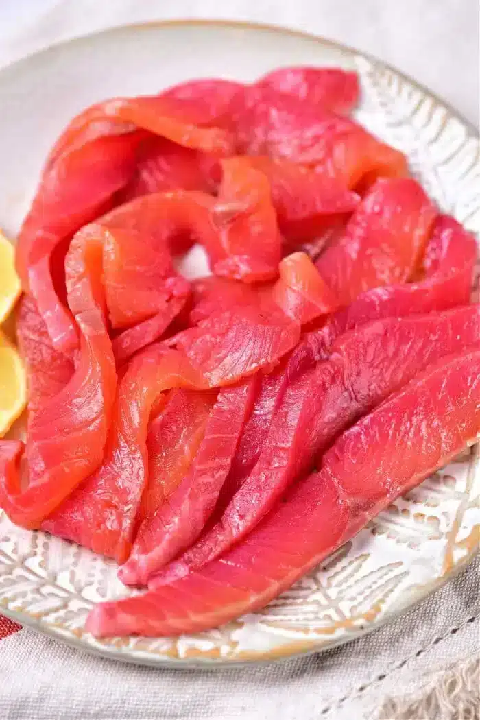 Slices of smoked salmon are artfully arranged on a white plate with lemon wedges on the side, offering a perfect presentation to showcase your skills in mastering how to cure salmon.