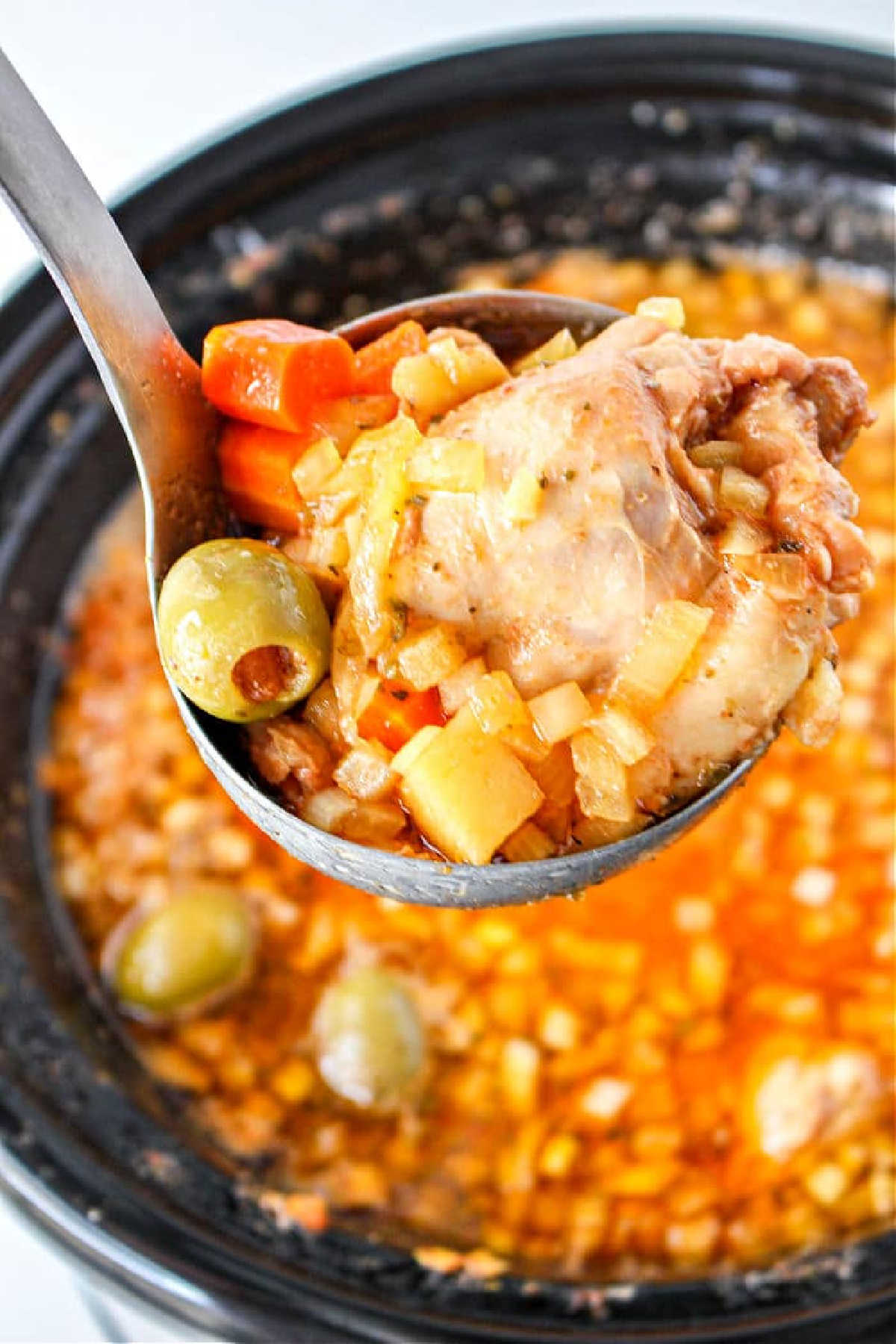 A ladle holds chicken, carrots, potatoes, and olives over a pot of Guisado de Pollo stew.