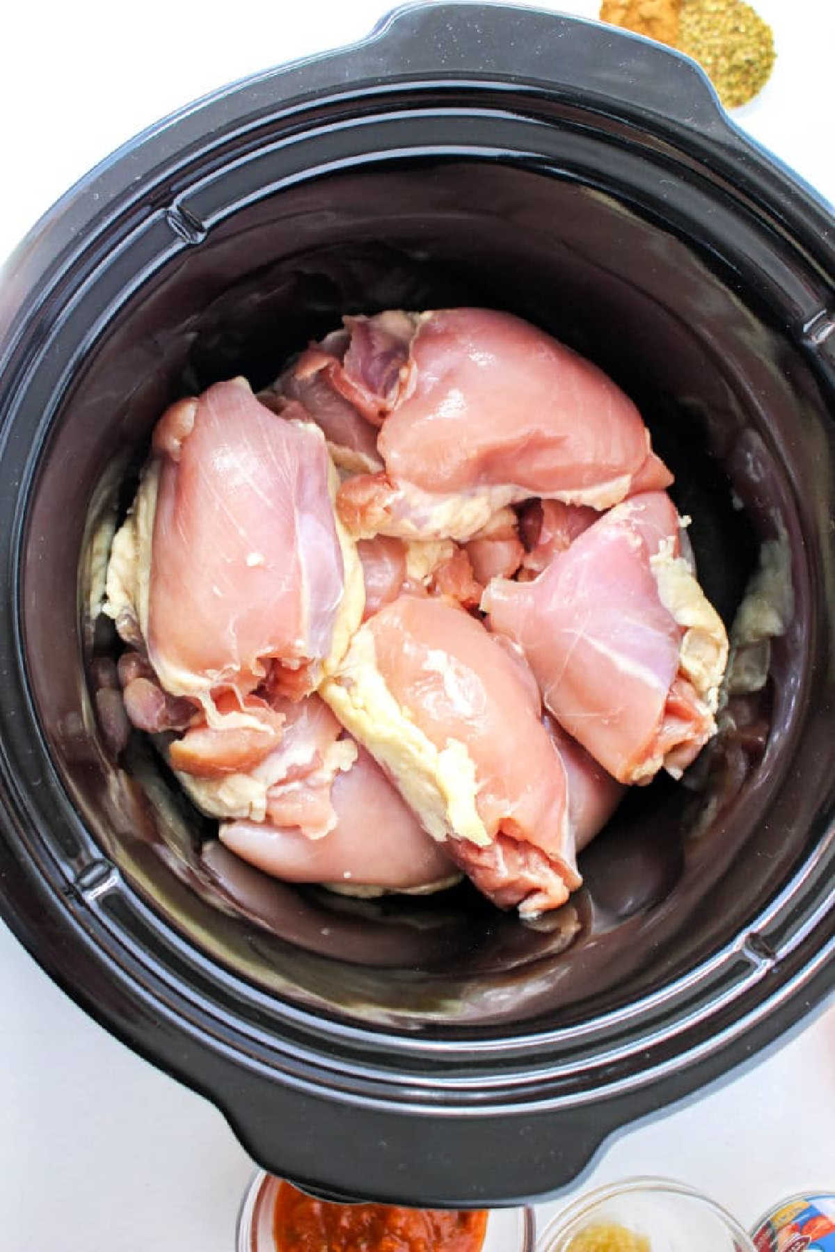 Raw chicken thighs arranged inside a black slow cooker, ready to be transformed into a flavorful Guisado de Pollo.
