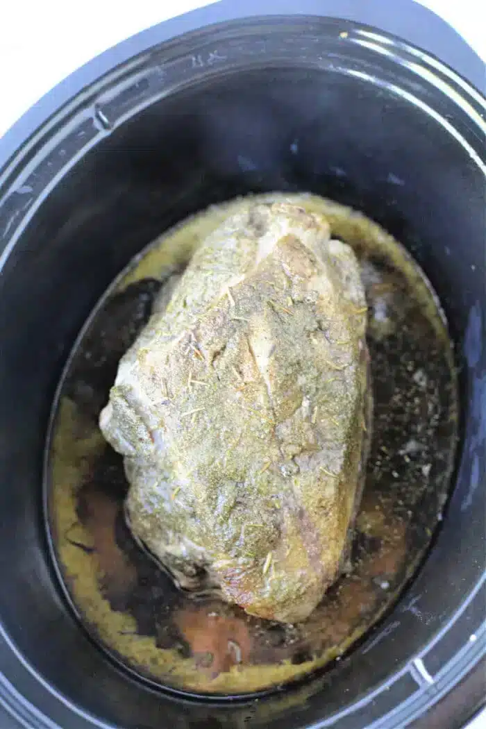 A black slow cooker contains a tender, 3 Ingredient Pork Roast Slow Cooker bone-in pork shoulder roast. The roast is seasoned with herbs and sits in its juices at the bottom of the cooker.