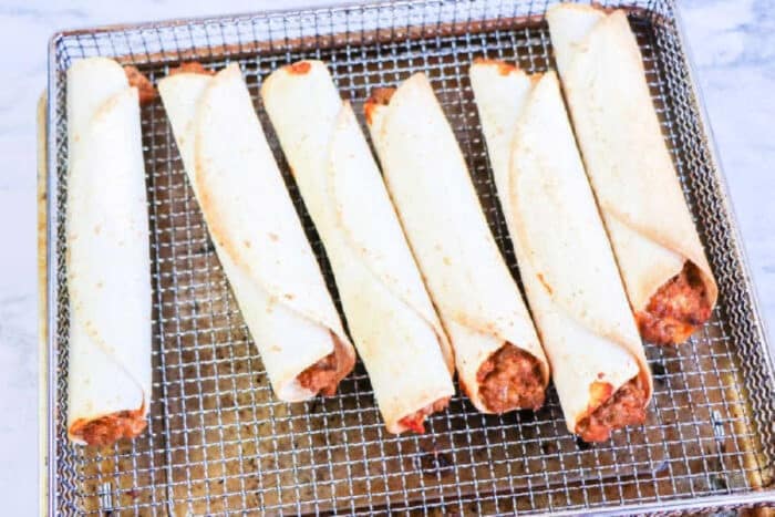 Six uncooked air fryer flautas filled with meat are arranged on a wire rack, ready to be cooked.
