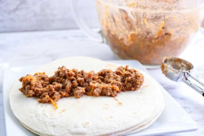 Air fryer flautas filled with seasoned ground meat are served on a plate, with a mixing bowl and scoop in the background.