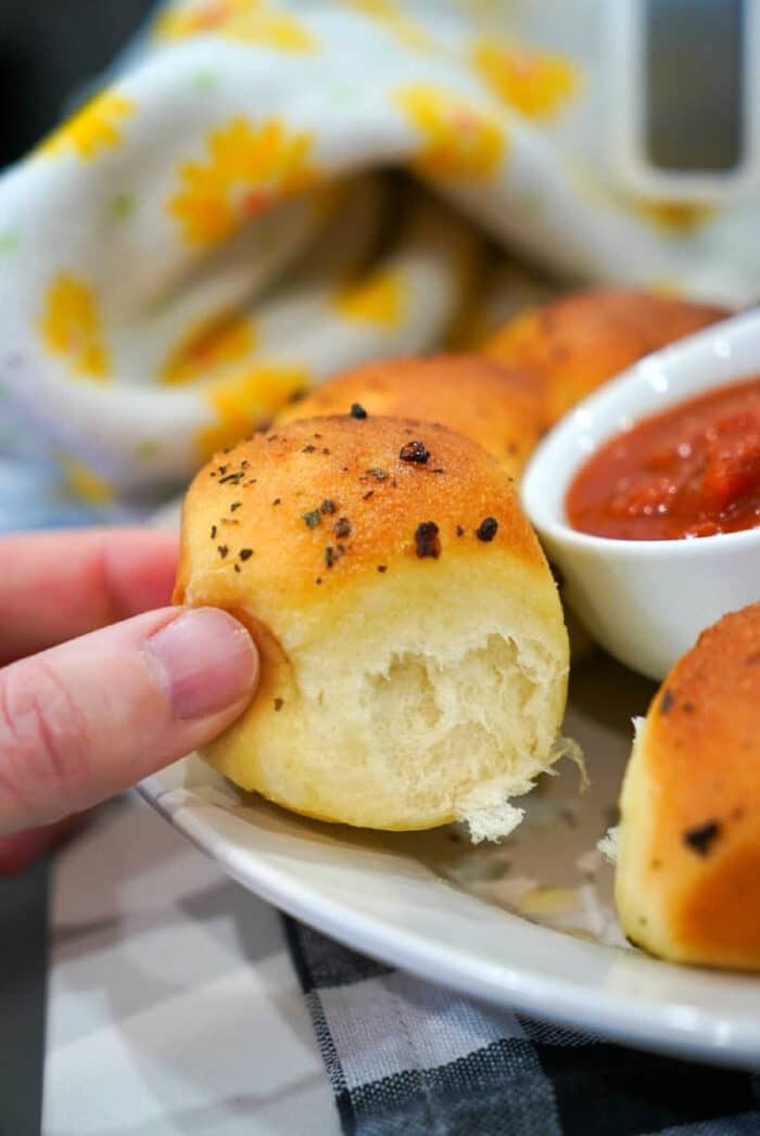 A hand holding a garlic knot roll, baked from Frozen Rolls in Air Fryer, next to a bowl of marinara sauce on a plate.