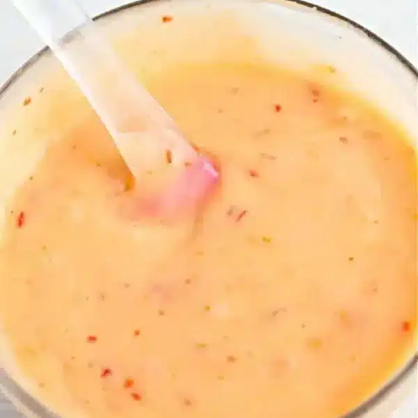 A clear bowl brimming with creamy bang bang shrimp sauce, its orange hue flecked with small red specks. A transparent spoon rests within, hinting it has just been stirred. The light, neutral background completes the inviting scene.