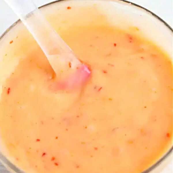 A glass bowl of creamy, orange bang bang shrimp sauce with red specks, stirred with a clear spoon.