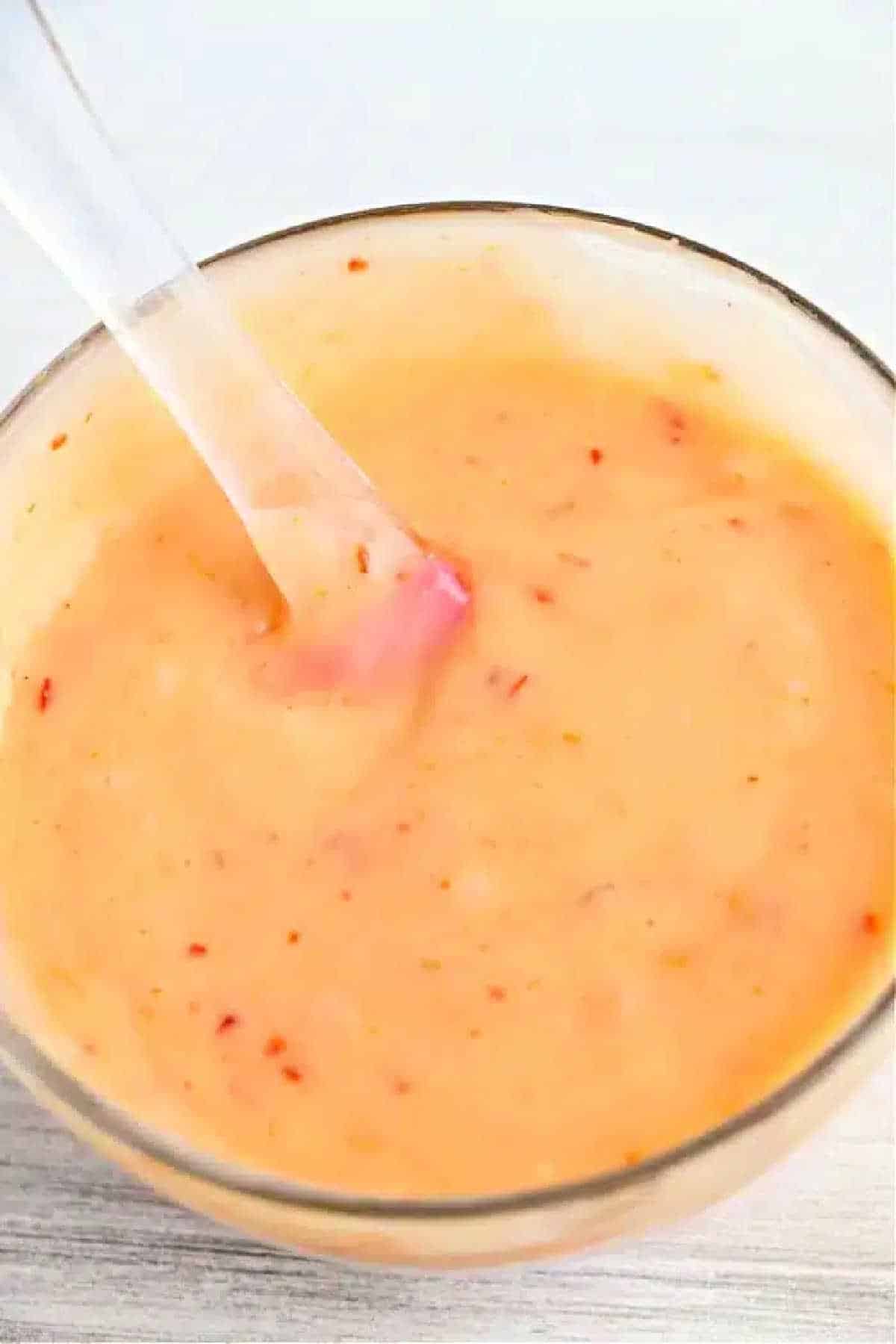 A glass bowl of creamy, orange bang bang shrimp sauce with red specks, stirred with a clear spoon.