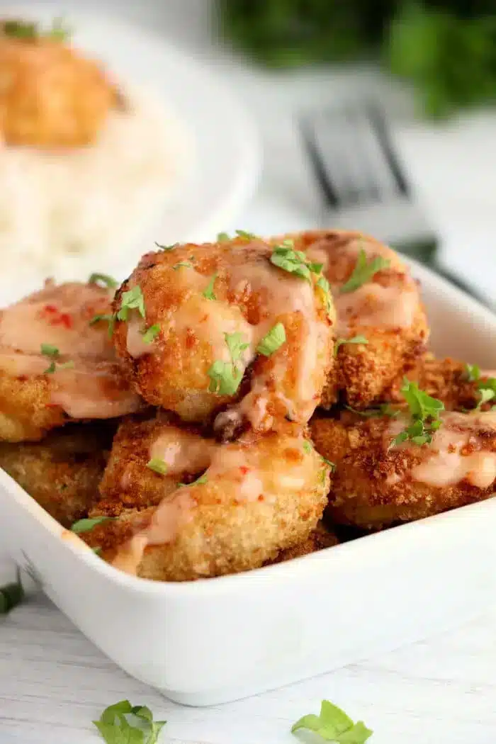 A rectangular white dish filled with crispy fried shrimp drizzled with bang bang shrimp sauce. The shrimp are garnished with chopped parsley. A fork and a plate of rice are partially visible in the background.