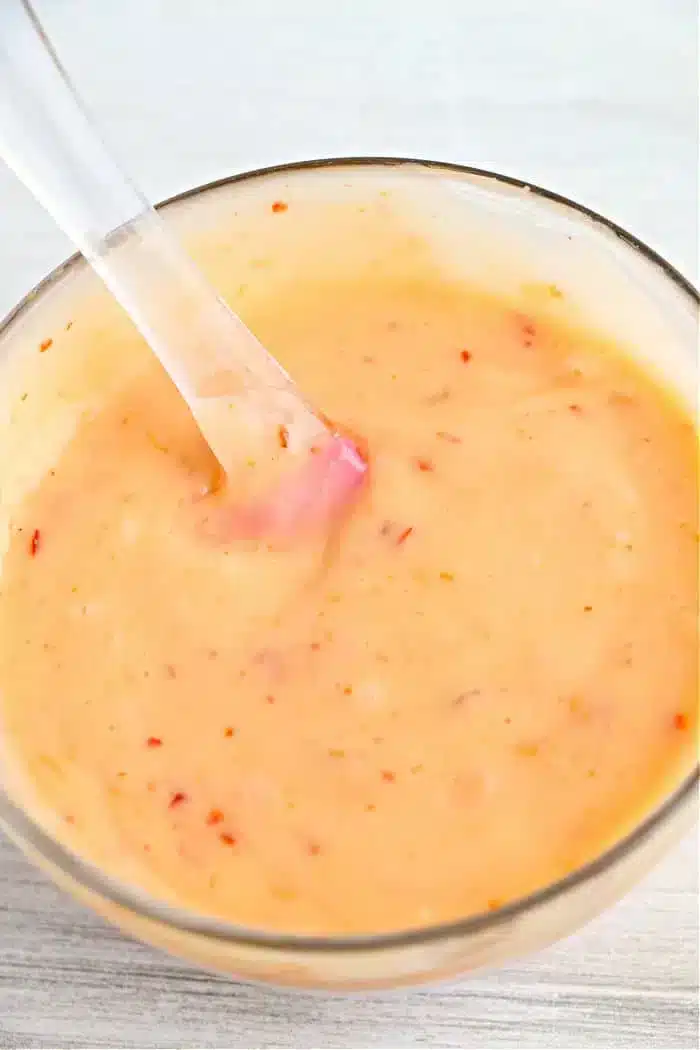 A glass bowl filled with a creamy, orange bang bang shrimp sauce spotted with small red specks. A clear spoon rests inside the bowl, ready to serve. The surface beneath is a light, textured material.