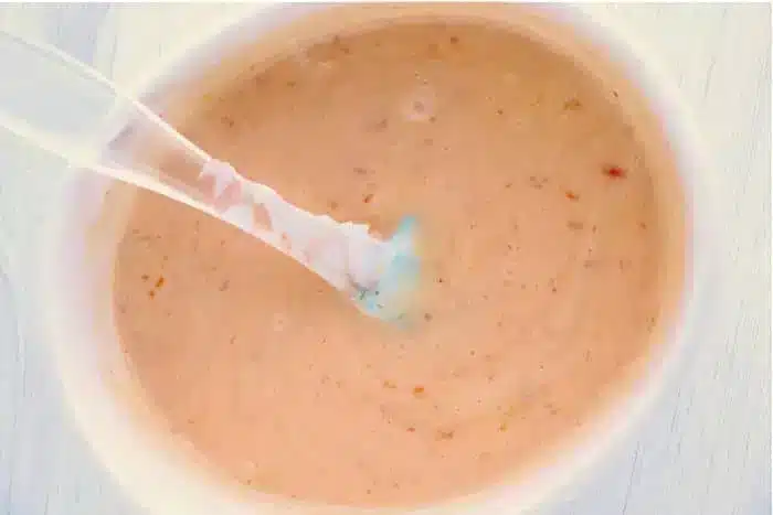 A plastic spoon is dipped into a container of creamy bang bang shrimp sauce, with visible flecks of seasoning throughout. The sauce has a light orange hue and a smooth texture, reminiscent of Thousand Island dressing.