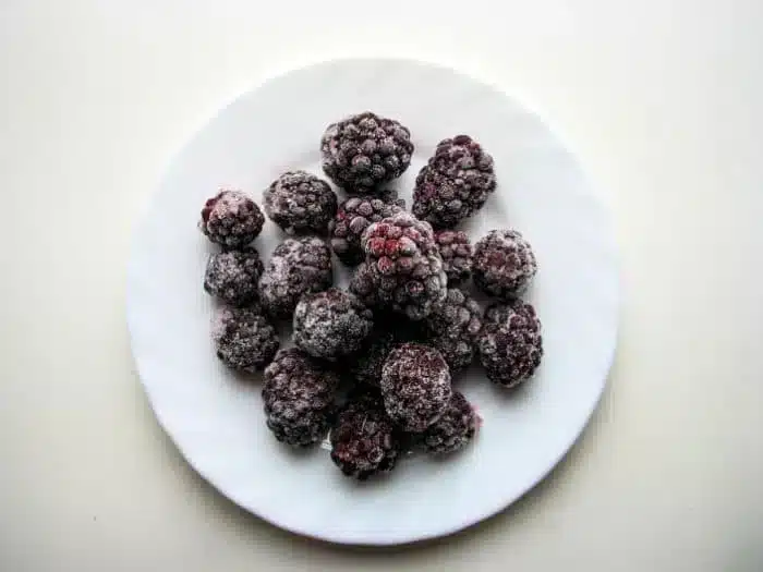 A white plate containing a small pile of frozen blackberries, perfect for crafting a delightful blackberry cobbler, is centered on a light background.