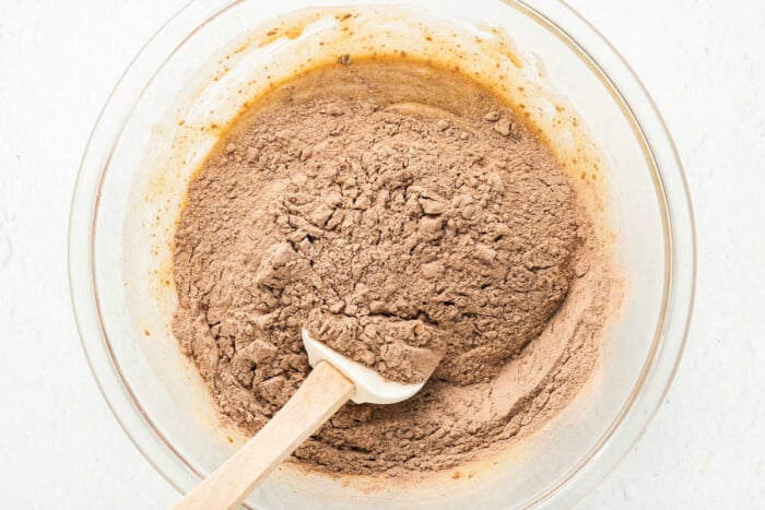 A glass bowl contains a mixed brown dry ingredient, likely cocoa powder and flour, with a white spatula resting inside. It seems to be the beginnings of cherry brownies, as the mixture is in the process of being stirred, indicating baking preparation.