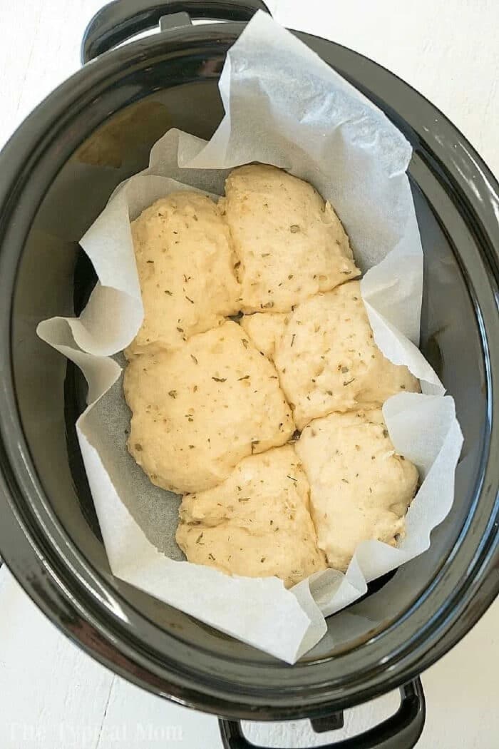 These easy slow cooker no yeast rolls are the perfect side dish with lasagna or any other pasta dish! There's no need to wait for them to rise and they cook by themselves right in your crockpot. Dripping with melted butter and garlic with a dash of parmesan they'll become your go to bread on busy evenings. #rolls #noyeast #easy #recipe #slowcooker #bread #roll #crockpot #garlic Unbaked herb biscuits, resembling Slow Cooker Rolls, are placed on parchment paper inside a black slow cooker, ready to be cooked.