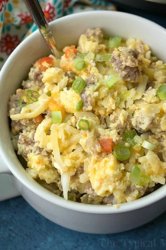 Bowl of scrambled eggs with sausage, cheese, potatoes, and chopped green onions, with a spoon—just like the comforting flavors you’d find in a classic crockpot breakfast casserole recipe.