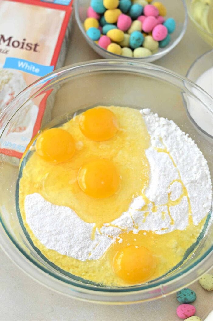 Four eggs and cake mix in a glass bowl, with an Easter Bundt Cake mix box and candy eggs in the background.