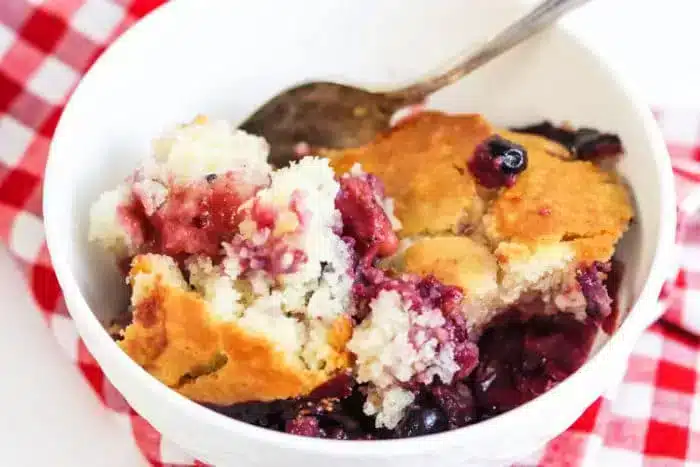 A white bowl holds a serving of blackberry cobbler with a golden crust made with Bisquick. A spoon is nestled in the bowl, which rests on a red and white checkered cloth.