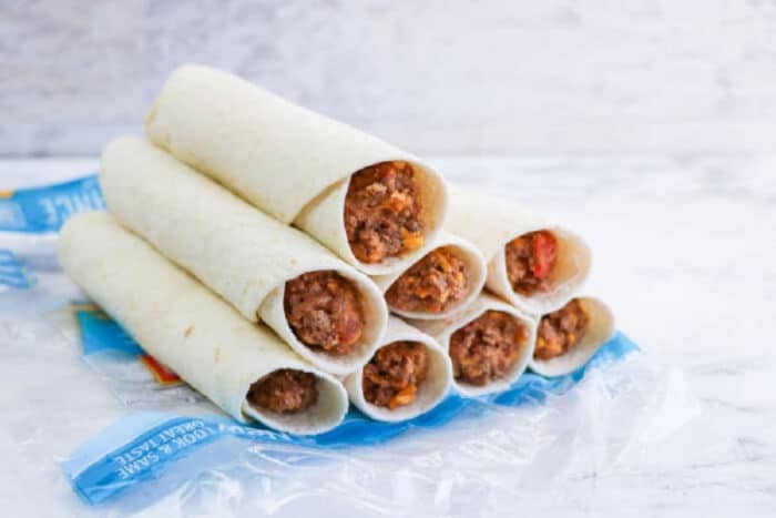 Eight air fryer flautas, filled with beef and cheese, are stacked on a blue plastic wrapper against a light background.