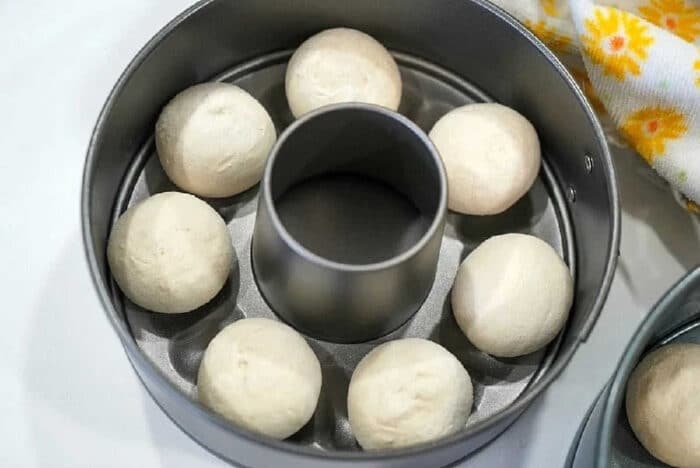 Eight dough balls, perfect for making Frozen Rolls in Air Fryer, are arranged in a ring mold pan and ready for baking on a white surface.