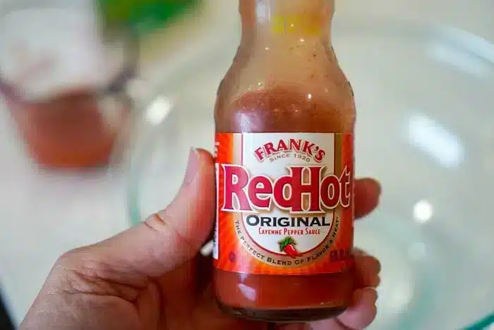 A hand holds a bottle of Frank's RedHot Original Cayenne Pepper Sauce with its signature red label. In the blurry background, a measuring cup and glass bowl hint at a delicious creation—perhaps the perfect bang bang shrimp sauce in the making.