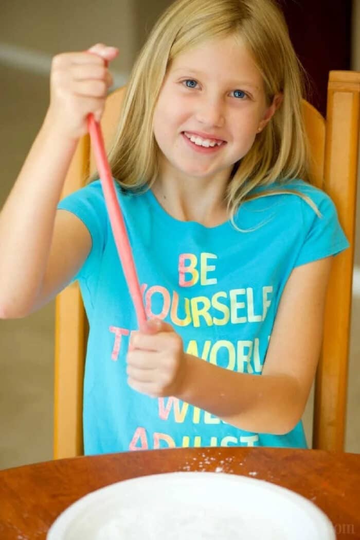 Edible slime recipe with only 3 ingredients! This is an easy and safe slime recipe that you can eat and tastes great. Use gummy bears for this! Best edible slime recipe ever. #edibleslime #slime #safe #boraxfree #noborax #gummybear #easy #best Smiling girl in a blue shirt stretches pink slime over a bowl while sitting at a table, showing how to make edible slime.