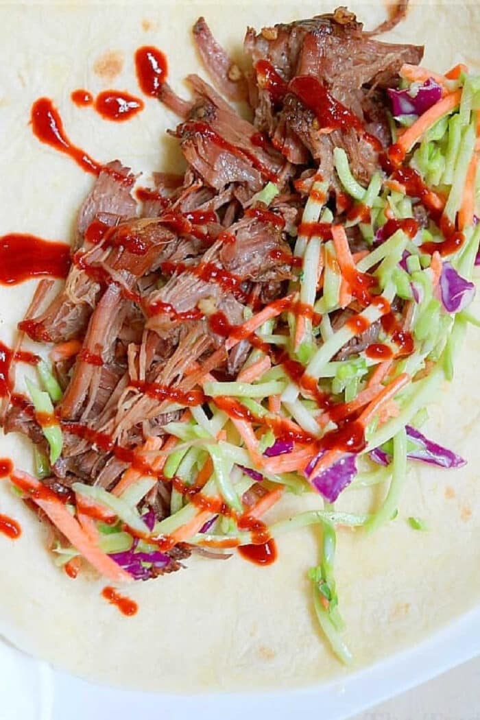 Slow cooker Korean beef, slaw, and red sauce on a tortilla, ready to be wrapped.