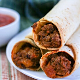 Three crispy ninja foodi air fryer flautas rest on a white plate with a side of salsa, placed on a striped cloth.