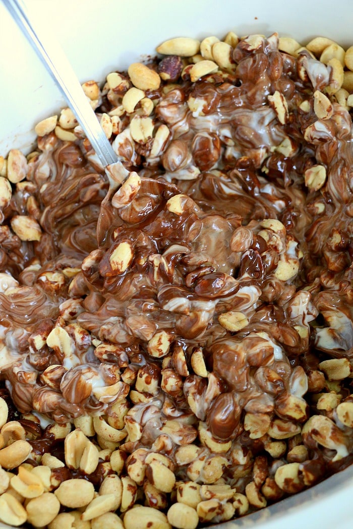 Close-up of crockpot peanut clusters, with peanuts mixed into melted chocolate and white cream, being stirred with a spoon.