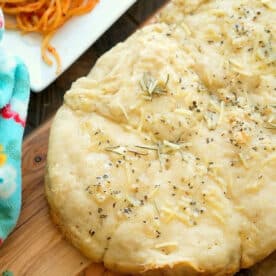 Homemade focaccia bread topped with herbs and cheese on a wooden board, with pasta in the background—perfect alongside warm Slow Cooker Rolls for a comforting meal.