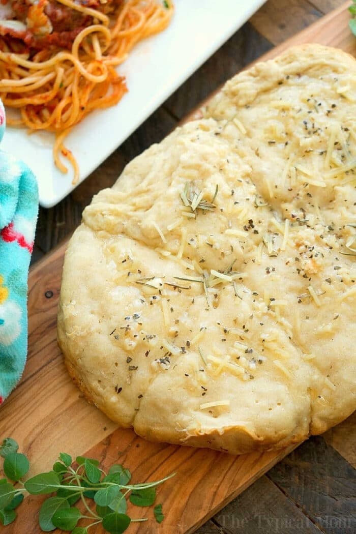 Slow cooker no yeast rolls are the perfect side dish with lasagna or any other pasta dish! There's no need to wait for them to rise and they cook by themselves right in your crockpot. Dripping with melted butter and garlic with a dash of parmesan they'll become your go to bread on busy evenings. #rolls #noyeast #easy #recipe #slowcooker #bread #roll #crockpot #garlic Homemade focaccia bread topped with herbs and cheese on a wooden board, with pasta in the background—perfect alongside warm Slow Cooker Rolls for a comforting meal.