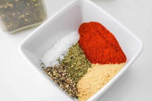 A square white bowl holds a vibrant array of spices—black pepper, salt, dried herbs, and ground red pepper—perfect for seasoning a Pot Roast Chicken. In the blurred background, a spice jar hints at the delicious possibilities.