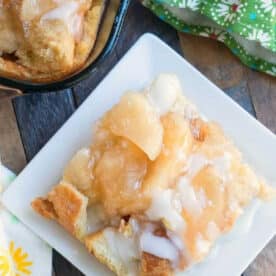 A square of apple pie bread pudding with glaze on a white plate, next to a baking dish and green patterned napkin.
