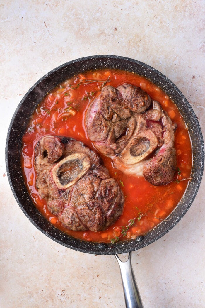 Two pieces of beef shank with bone simmering in tomato sauce in a black skillet on a light countertop.