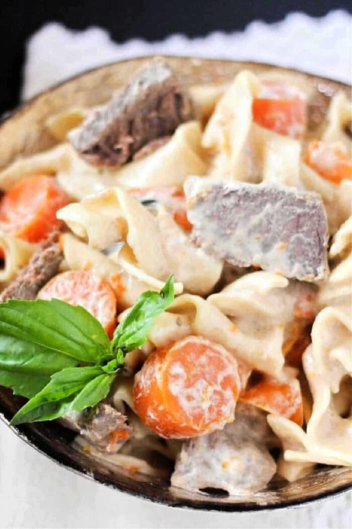 A bowl of creamy crockpot beef stroganoff and noodle stew with carrots, garnished with fresh basil.