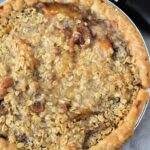 A freshly baked canned peach pie with a crumbly oat topping sits enticingly next to an ice cream scooper.