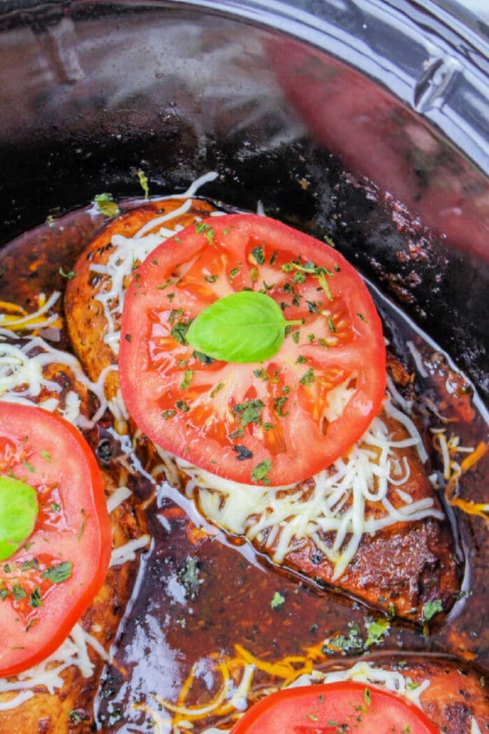 Crockpot Chicken Frozen Breasts topped with melted cheese, tomato slices, and basil simmer in a slow cooker with savory sauce.
