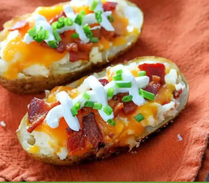 Make Ahead Twice Baked Potatoes: Two baked potato halves topped with melted cheese, crispy bacon pieces, sour cream, and chopped green onions are beautifully placed on an orange cloth.