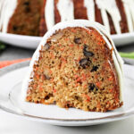 A slice of carrot pound cake with nuts and white icing graces the plate, while the whole cake sits enticingly in the background.