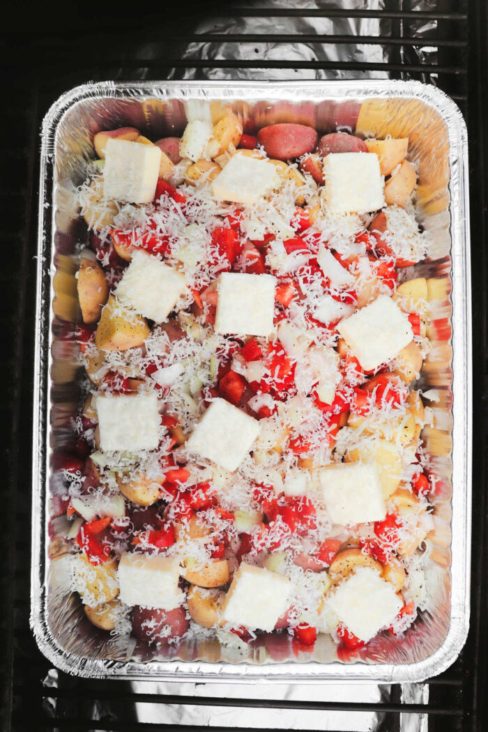 Foil pan filled with chopped vegetables, grated cheese, and cubed butter, perfect for making smoked cheesy potatoes and ready to be cooked.