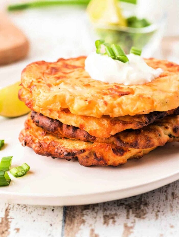 Three cheesy air fryer potato pancakes stacked on a plate, topped with sour cream and chopped green onions.