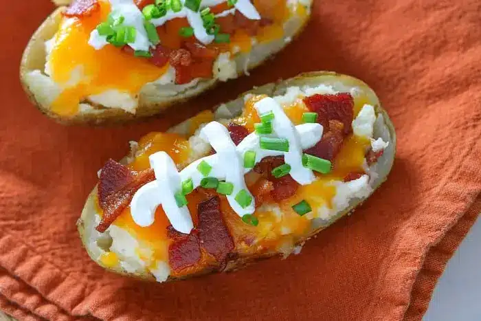 Two loaded baked potato halves rest on an orange cloth, each topped with sour cream, chopped bacon, melted cheddar cheese, and green onions. Perfect for make-ahead twice-baked potatoes, these savory delights are sure to impress at any meal.