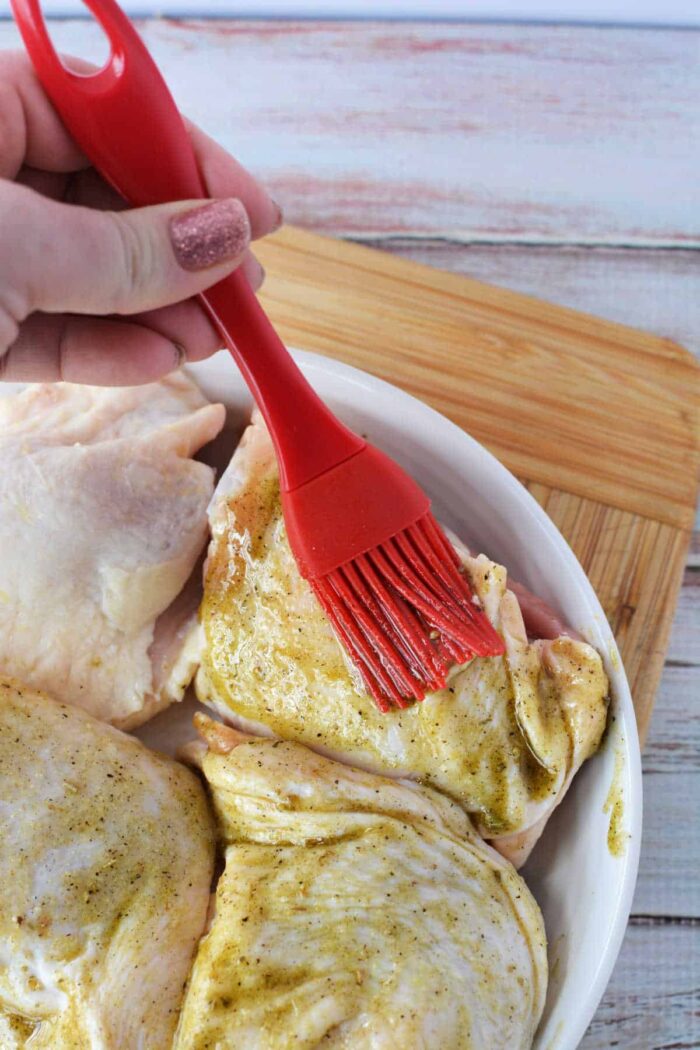 A hand uses a red brush to spread marinade on raw chicken pieces in a white dish, prepping them before discovering how long to bake chicken thighs at 425 for perfect results.