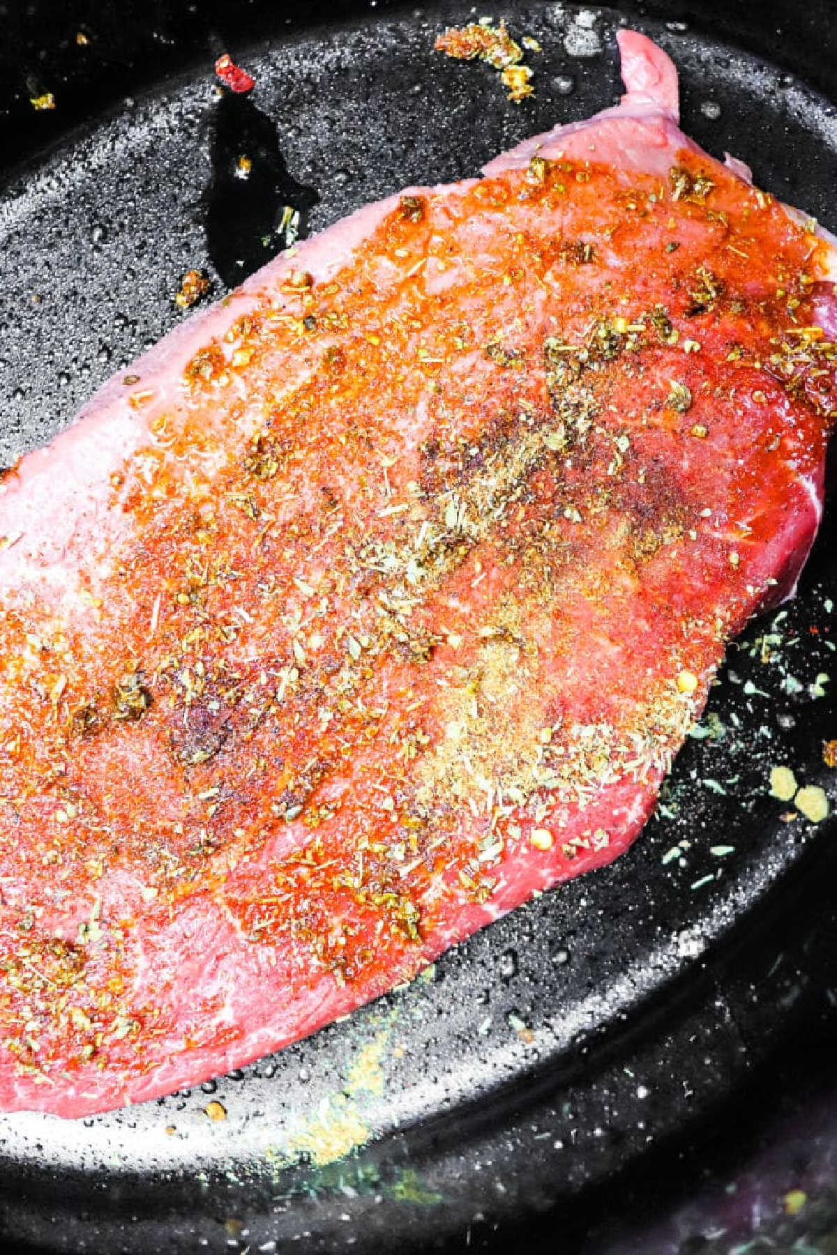 Raw seasoned beef steak on a black surface, sprinkled with various spices and herbs, perfect for making Carne Asada Crock Pot recipes.