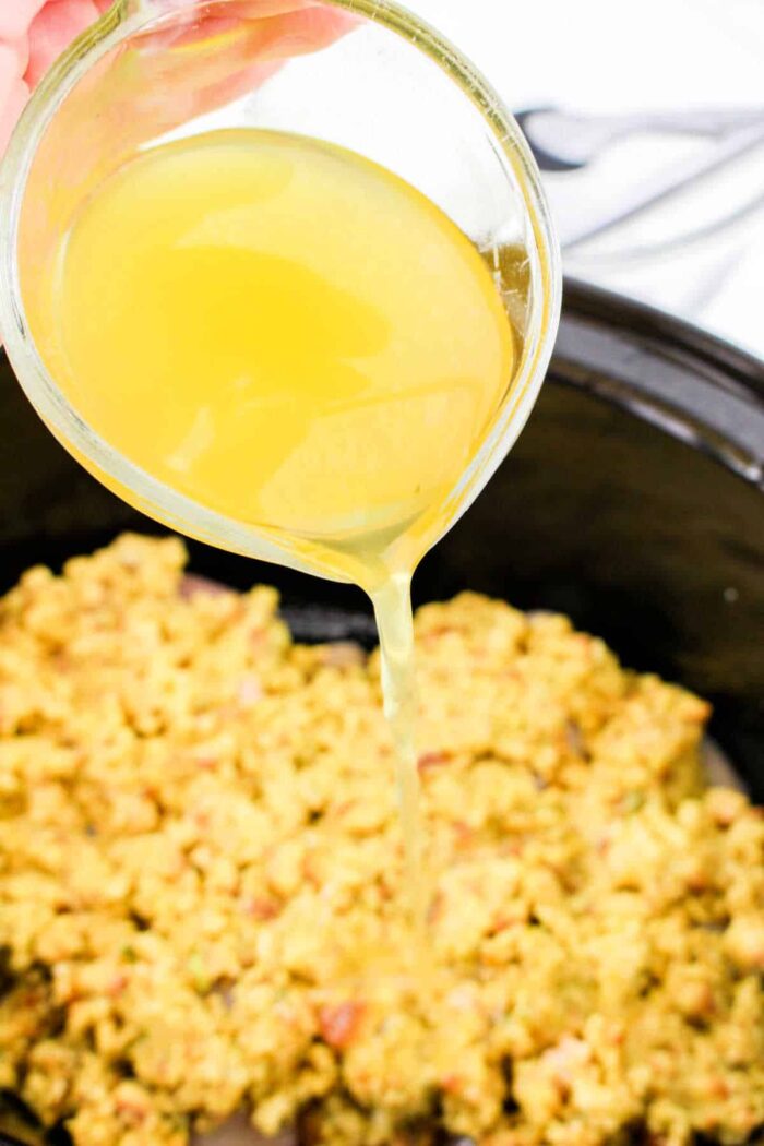 A hand pours broth from a glass measuring cup over Chicken and Stuffing Crockpot mix in a slow cooker.