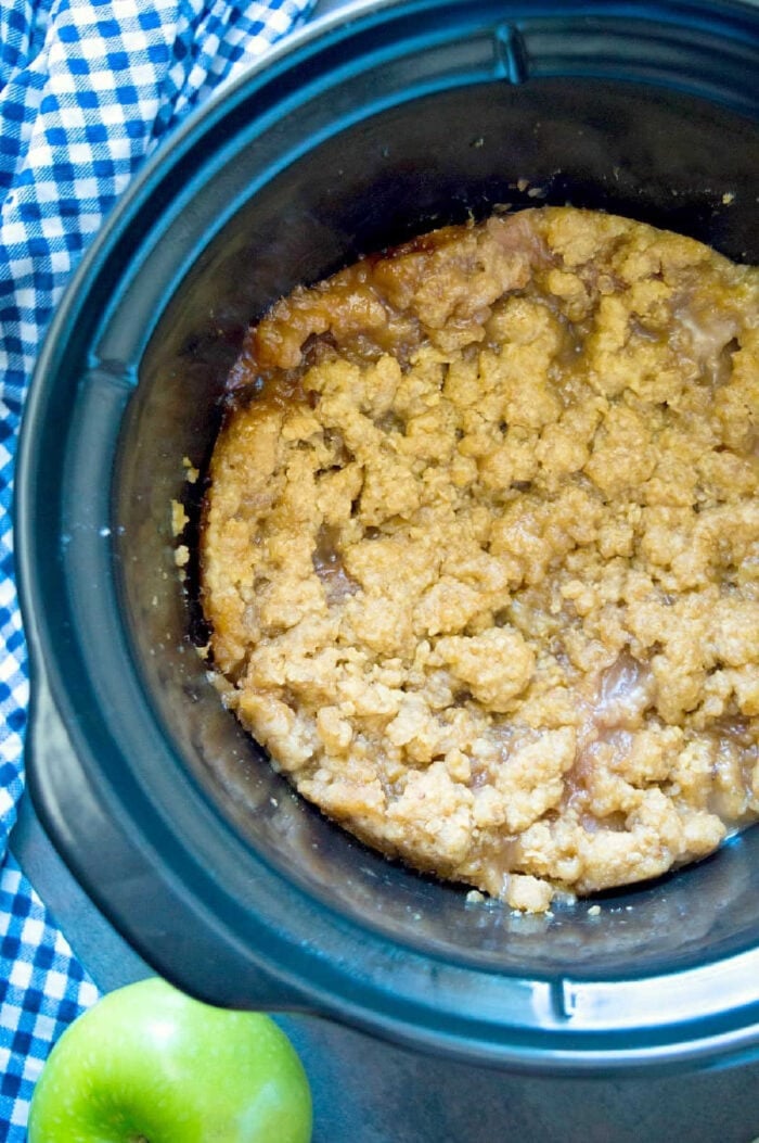 Crockpot apple crisp pie is amazing! It tastes like the warm apple pie filling without the crust and cooked in your slow cooker, easy! #applecrisp #crockpot #slowcooker #pie #dessert #fruit #breakfast #crockpotrecipes #crockpotapplecrisp #thetypicalmom A crockpot filled with warm crockpot apple crisp sits beside a green apple and a blue checkered cloth.